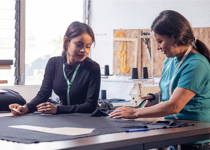 Two women collaborating on fabric cutting at a sewing workshop with a sewing machine in the background.