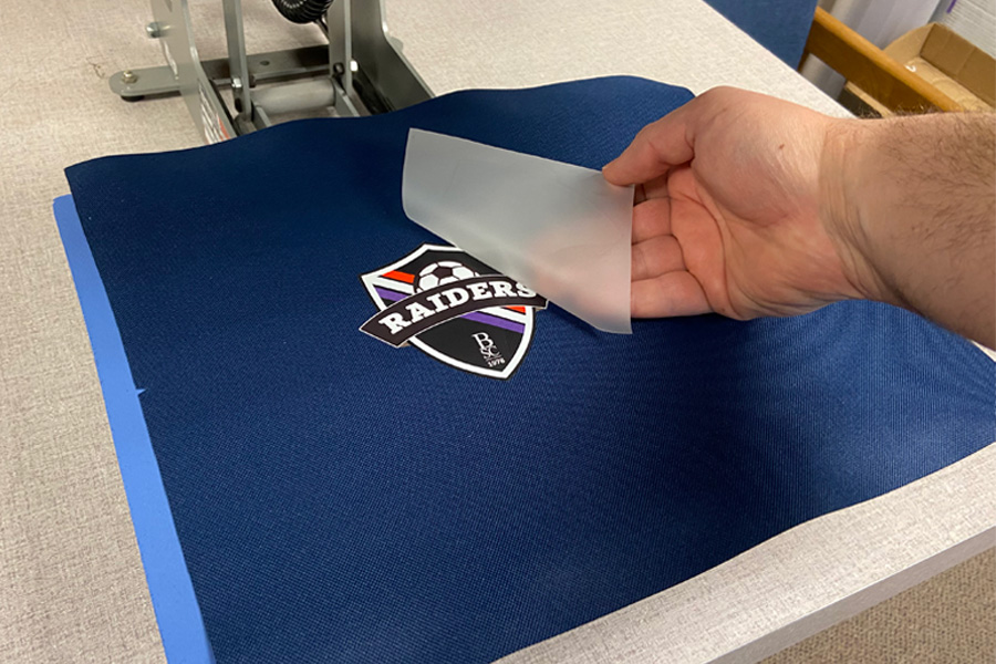 A hand holding a transparent sheet over a navy fabric with a Raiders logo on it.